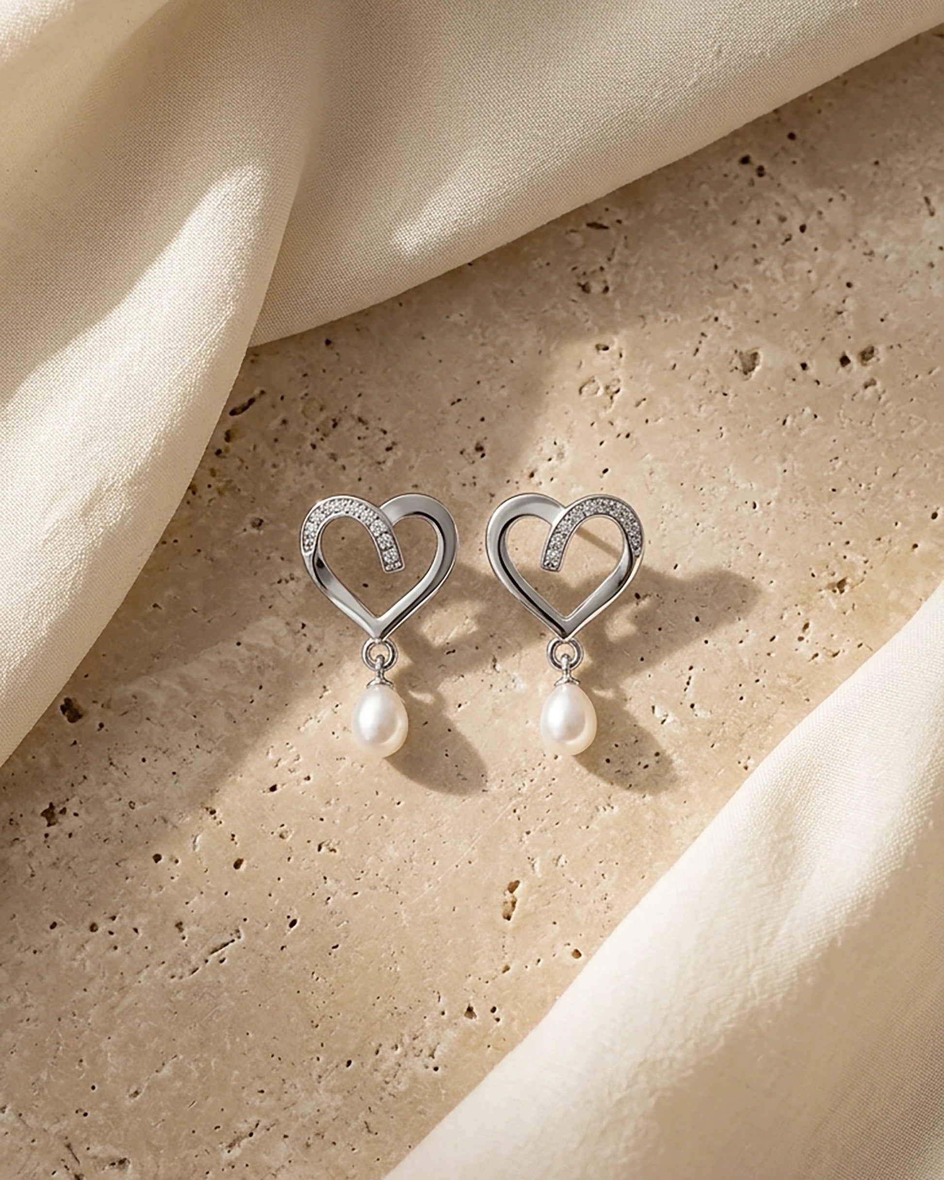 Silver heart and pearl earrings styled on a neutral stone surface with soft draped fabric